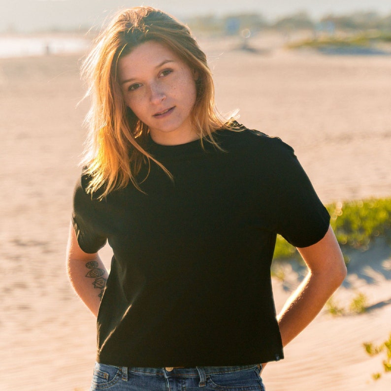 K&ouml;nnte beinhalten: Ein schwarzes T-Shirt mit Rundhalsausschnitt. Das kurz&auml;rmelige Shirt wird mit einer blauen Jeans getragen. Die Person auf dem Bild hat rotes Haar und steht an einem Strand. Der Hintergrund ist unscharf.