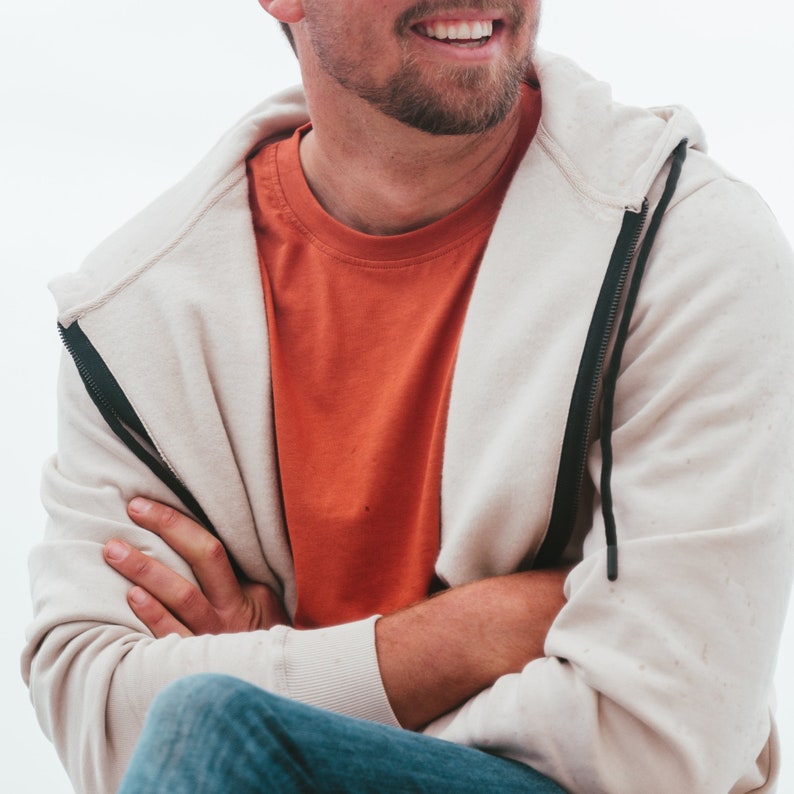 K&ouml;nnte beinhalten: Ein hellbeige Rei&szlig;verschluss-Hoodie mit schwarzem Rei&szlig;verschluss und einem orangefarbenen Rundhals-T-Shirt. Die Person tr&auml;gt eine blaue Jeans. Der Hoodie hat eine Kapuze und einen Kordelzug. Die Person hat die Arme verschr&auml;nkt.
