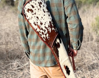 Peut inclure: Un étui à fusil en peau de vache avec une garniture et une sangle en cuir marron. L'étui présente un fond blanc avec des taches marron et un bord tressé. L'étui est porté sur l'épaule.
