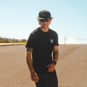 May include: Black t-shirt with a small white graphic on the chest, worn with a black cap and sunglasses. The cap has a brown patch on the front. The person is standing on a paved road under a clear blue sky.