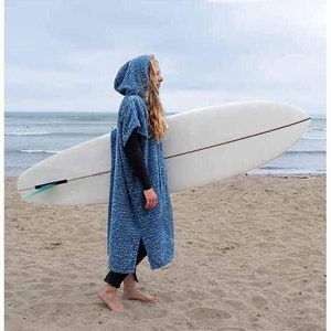 May include: A person wearing a blue hooded towel robe with a white surfboard under their arm. The person is standing on a sandy beach.