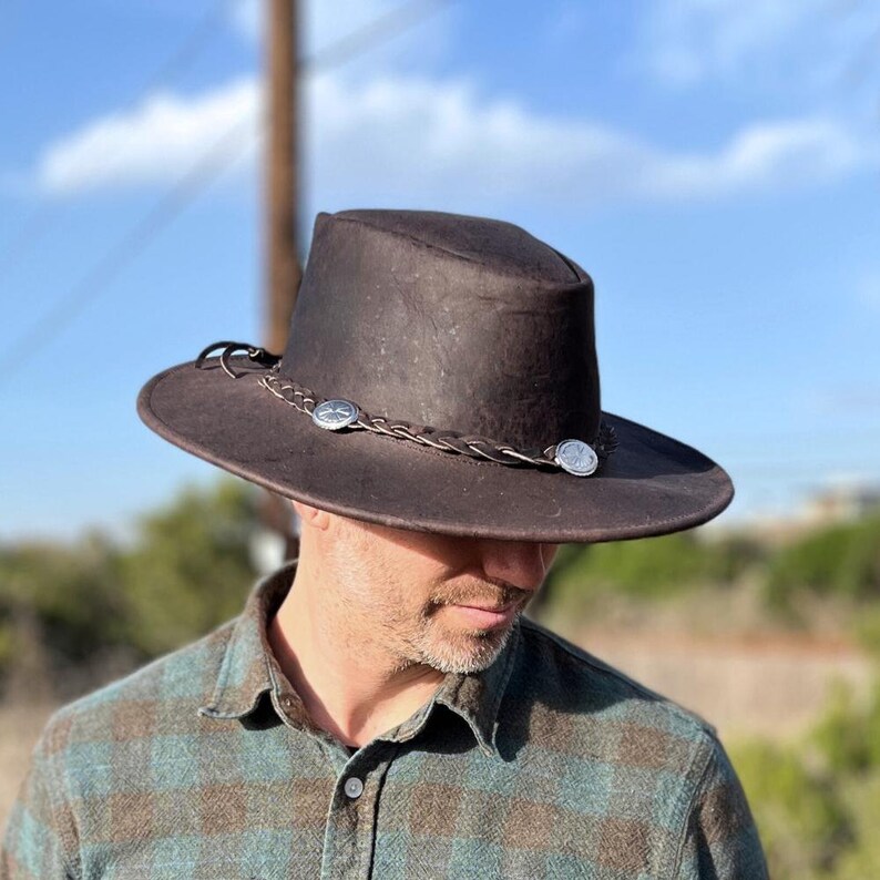 May include: A brown leather cowboy hat with a braided band and silver accents. The hat is worn by a person with a beard and a plaid shirt.