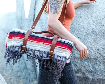 May include: A rolled-up blanket with red, black, and white stripes, secured with brown leather straps and a handle. The blanket has fringed edges and is carried over the shoulder. The straps have gold-colored buckles.