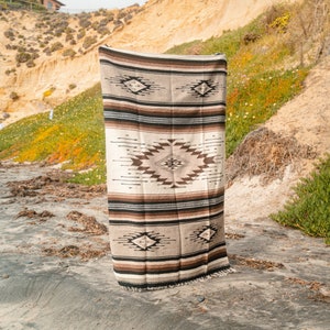 May include: A brown and white woven blanket with a geometric pattern. The blanket is hanging on a sandy beach.