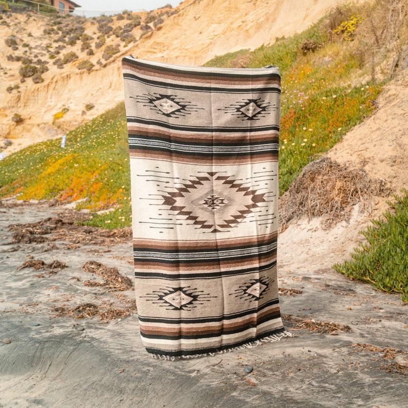 May include: A brown and white woven blanket with a geometric pattern. The blanket is hanging on a sandy beach.