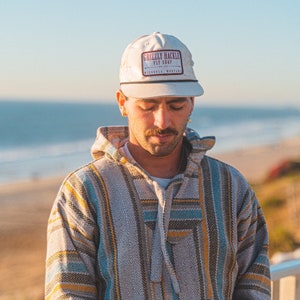 May include: A white cap with a rectangular patch that reads "GRIZZLY HACKLE FLY SHOP" in maroon lettering. The cap is worn with a patterned hoodie in shades of gray, blue, and yellow. The background shows a beach and ocean.