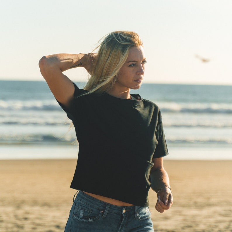 K&ouml;nnte beinhalten: Ein schwarzes kurz&auml;rmeliges T-Shirt und blaue Jeans. Die Person steht an einem Sandstrand mit dem Meer im Hintergrund. Die Person h&auml;lt sich die Haare mit der linken Hand.