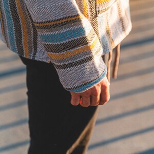May include: A close-up of a woven jacket with a herringbone pattern. The jacket features vertical stripes in shades of gray, yellow, blue, and black. The sleeve cuff is trimmed with blue. The person is wearing black pants.