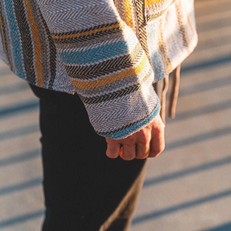 May include: A close-up of a woven jacket with a herringbone pattern. The jacket features vertical stripes in shades of gray, yellow, blue, and black. The sleeve cuff is trimmed with blue. The person is wearing black pants.
