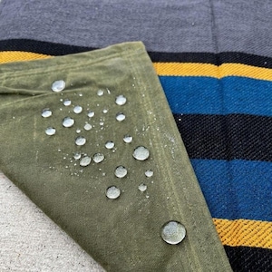 May include: Close-up of a green, water-resistant fabric with water droplets. The fabric is folded over a striped textile with black, blue, and yellow stripes. The background is a solid gray fabric.