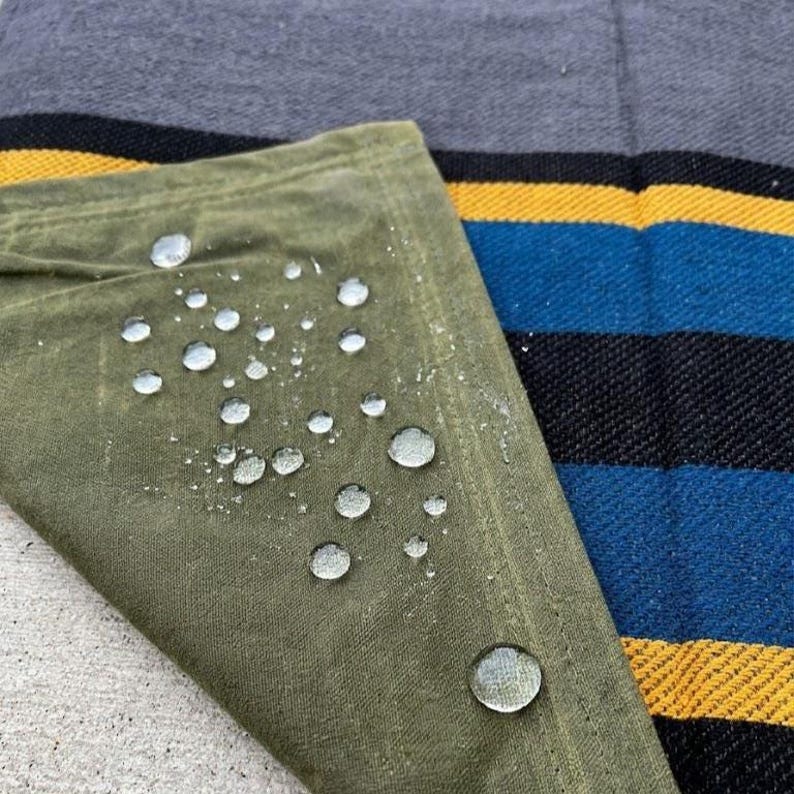 May include: Close-up of a green, water-resistant fabric with water droplets. The fabric is folded over a striped textile with black, blue, and yellow stripes. The background is a solid gray fabric.