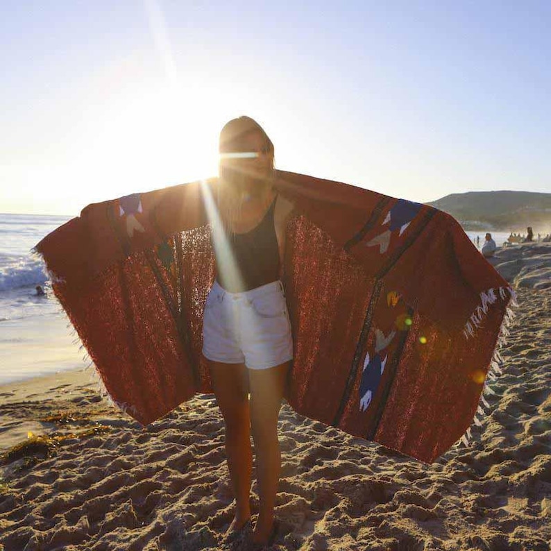 May include: A person wearing a white tank top and white denim shorts is standing on a sandy beach. They are holding a large, red and blue patterned blanket over their shoulders.