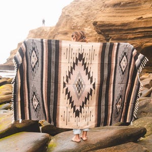 May include: A person standing on a rocky beach holding a large, woven blanket with a black, white and brown geometric pattern.