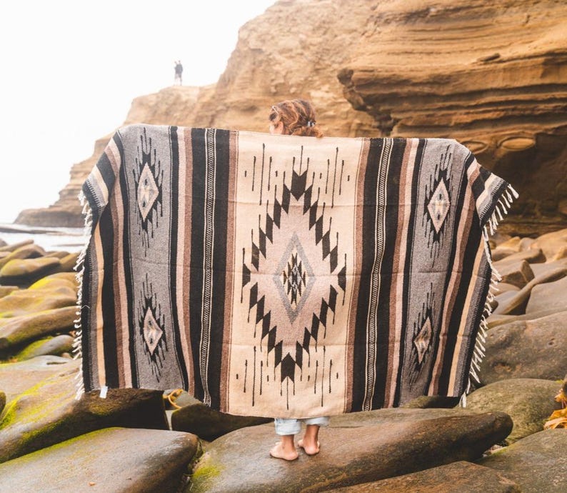 May include: A person standing on a rocky beach holding a large, woven blanket with a black, white and brown geometric pattern.