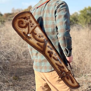 Op de afbeelding: Een bruine leren geweerkoffer met ingewikkelde bloemen- en krulwerkontwerpen. De koffer heeft een schouderriem en gespsluitingen. Het leer is een rijke, warme bruine kleur en het ontwerp is in reliëf.