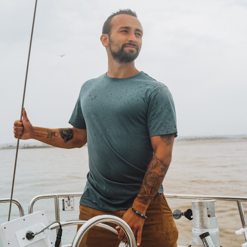 May include: A teal short-sleeved t-shirt and brown trousers are worn on a boat. The person is holding a rope and looking off into the distance. The person has tattoos on their arms and a bracelet on their wrist. The sky is overcast.
