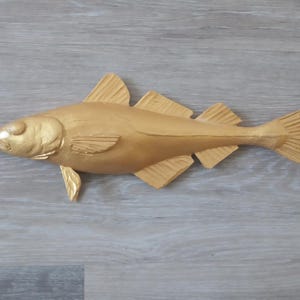 Pode incluir: Escultura de peixe dourado esculpida com barbatanas e cauda detalhadas. O peixe está disposto horizontalmente, mostrando sua forma e textura. A escultura é provavelmente feita de madeira ou material semelhante.