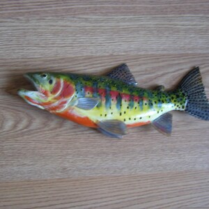 Puede incluir: Una figura de pez pintada y colorida con la boca abierta, sobre una superficie de madera. El pez presenta una vibrante paleta de colores rojos, amarillos, verdes y azules, con manchas negras y aletas detalladas. El pez mide unos 30 cm de largo.