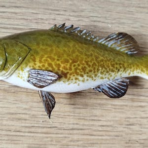 Pode incluir: Figura de peixe pintada com corpo amarelo-esverdeado, barriga branca e padrão salpicado. As barbatanas são brancas e pretas, e o peixe está sobre uma superfície de madeira. É um objeto decorativo.