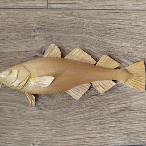 May include: A carved wooden fish sculpture, featuring a light brown color. The fish has detailed fins and a tail, with visible carved lines. The sculpture is lying on a wooden surface.