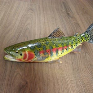 May include: A detailed, hand-painted fish sculpture. The fish has a green back, yellow belly, and red and yellow markings. The tail and fins are speckled with black and yellow dots. The sculpture is lying on a wooden surface, with the fish's mouth slightly open.