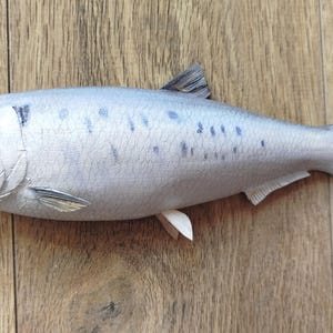 May include: A detailed, handcrafted fish sculpture with a light gray body and dark gray markings. The fish has a realistic design, including fins and a tail, and is set against a wooden background.