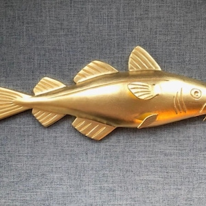 May include: A gold-colored fish wall ornament featuring a detailed molded body with defined scales, fins, and a tail. The fish is depicted in a horizontal swimming position with a visible eye, gill slits, and an open mouth, showcasing a smooth finish.