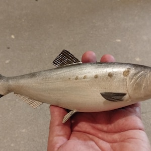 May include: A silver-toned fish figurine with a dark gray dorsal fin, tail, and pectoral fin. The body features a smooth, metallic finish with a row of small, circular indentations along the upper side and a clearly defined eye and mouth.