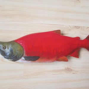 May include: A vibrant, hand-carved wooden fish sculpture. The fish features a bright red body, a green and black head, and a white mouth. The fins and tail are a reddish-brown color. The sculpture is displayed on a light-colored wooden surface.