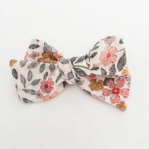 May include: A white fabric bow with a floral pattern in shades of pink, orange, and green. The bow is tied in a knot and has a simple design.