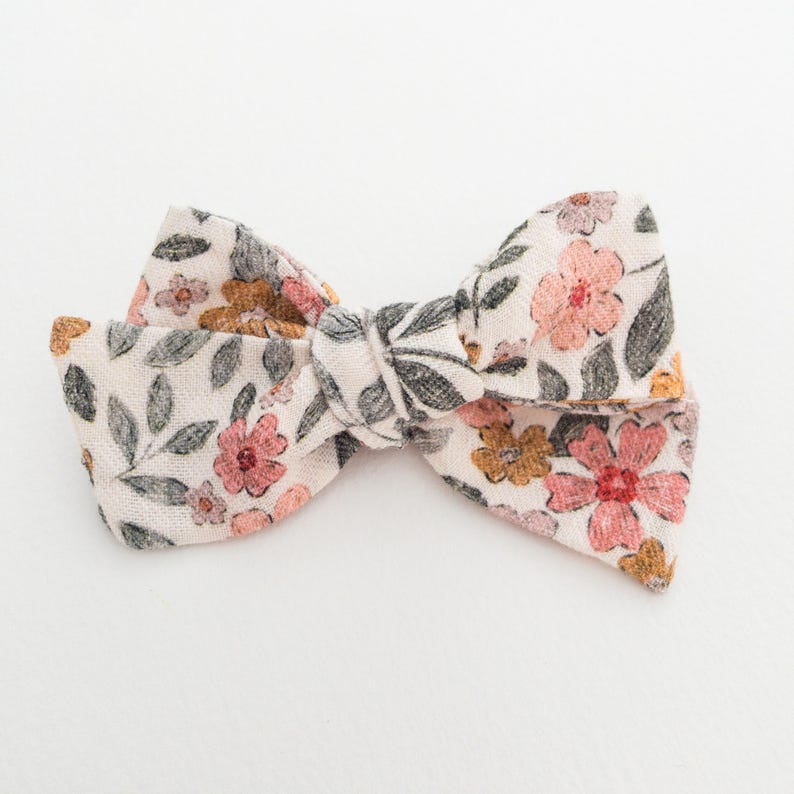May include: A white fabric bow with a floral pattern in shades of pink, orange, and green. The bow is tied in a knot and has a simple design.