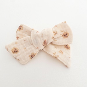 May include: A beige fabric hair bow with a floral pattern in brown. The bow is tied in a knot and has a soft, textured look.