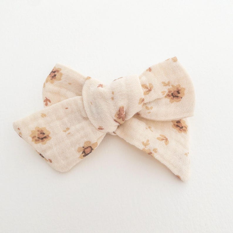 May include: A beige fabric hair bow with a floral pattern in brown. The bow is tied in a knot and has a soft, textured look.