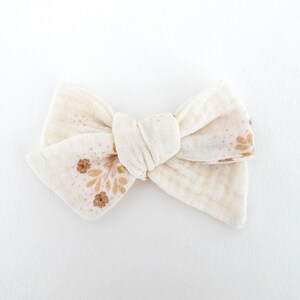 May include: A white fabric bow with a floral pattern in shades of pink and brown. The bow is tied in a simple knot and has a slightly textured surface.