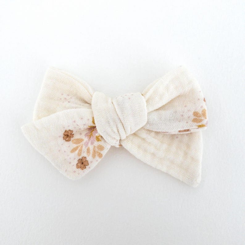 May include: A white fabric bow with a floral pattern in shades of pink and brown. The bow is tied in a simple knot and has a slightly textured surface.