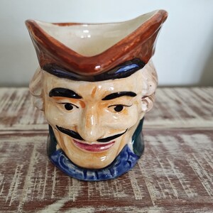 Könnte beinhalten: Ein Keramik-Krug in Form eines Männerkopfes mit einem braunen Hut. Der Krug hat einen blauen und grünen Kragen sowie einen gemalten Schnurrbart und Augenbrauen.
