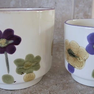 May include: Two white ceramic flower pots with purple and yellow floral designs. The larger pot is taller and has a wider base than the smaller pot.