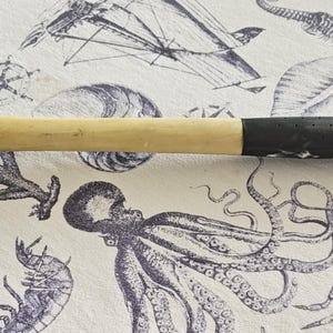 May include: A claw hammer with a wooden handle and a black rubber grip. The hammerhead is metal and shows signs of wear. The hammer is resting on a white cloth with a nautical design featuring illustrations of sea creatures and nautical elements.
