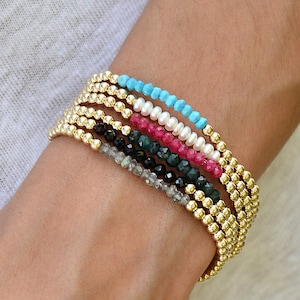 Può includere: Un set di braccialetti con perline dorate e pietre preziose di diversi colori, tra cui turchese, perle bianche, rosa, verde, nero e grigio.
