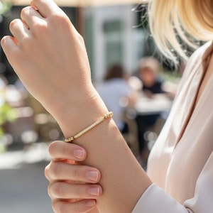 May include: A gold-toned bracelet featuring a central bar element encrusted with small, clear, sparkling stones, flanked by a series of small, round, polished gold-toned beads on either side, creating a delicate and refined wrist accessory.