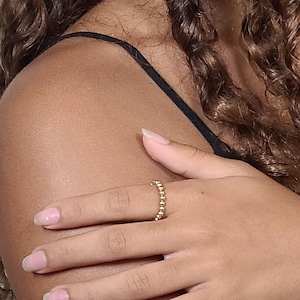 May include: A close-up of a person's hand wearing a gold beaded ring. The ring is made of small, round beads that are connected together to form a band. The ring is on the person's middle finger.
