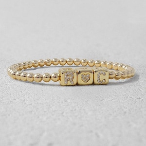 May include: Gold beaded bracelet with three gold square beads. The beads spell out "ROC" with the letter "R" encrusted with small clear stones, a heart, and the letter "C" also encrusted with small clear stones. The bracelet is on a white surface.