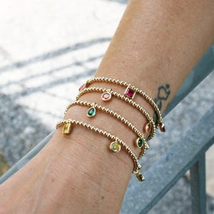 May include: A set of gold beaded bracelets with colorful gemstone charms. The charms are in the shapes of squares and teardrops.