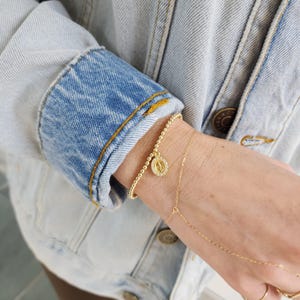 May include: A gold beaded bracelet with a small charm featuring a letter 'M' surrounded by small clear stones. The bracelet is worn on a person's wrist with a delicate gold chain bracelet on the other wrist.