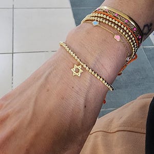 May include: A gold bracelet with a star of David charm. The bracelet is made of small gold beads and is worn on a person's wrist. The bracelet is stacked with other bracelets, including a gold chain bracelet with a pink heart charm and a gold chain bracelet with a blue heart charm.