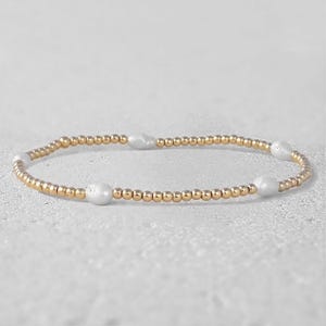 May include: A stretch bracelet composed of small, polished gold-toned spherical beads interspersed with four irregular, white, organic-shaped pearls arranged at intervals along the circular band of the jewelry piece.