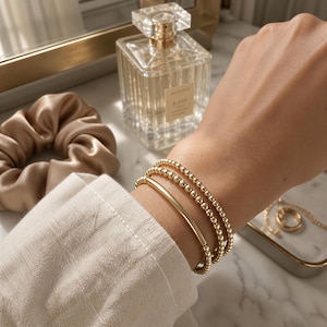 May include: A set of three gold-toned bracelets worn on the wrist. Two bracelets consist of small, uniform round beads, while the third bracelet features a smooth, elongated cylindrical bar. The jewelry is worn with a beige long-sleeved shirt.