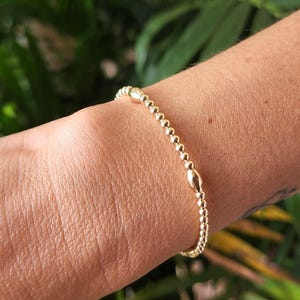 May include: A gold beaded bracelet with oval-shaped beads. The bracelet is on a person's wrist.