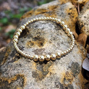 May include: A gold beaded bracelet with larger beads in the center. The bracelet is on a rough, brown, textured surface.