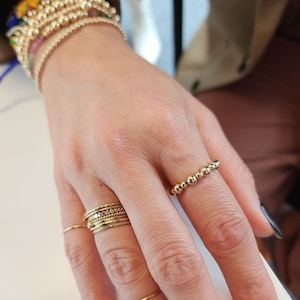 May include: A hand wearing a stack of gold rings on the middle finger and a single gold beaded ring on the ring finger.